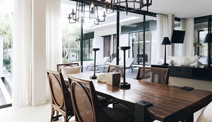 Elegant indoor dining area with a long wooden table and six rattan chairs, featuring modern black cage chandeliers and floor-to-ceiling glass overlooking the pool at Ambergris Villa