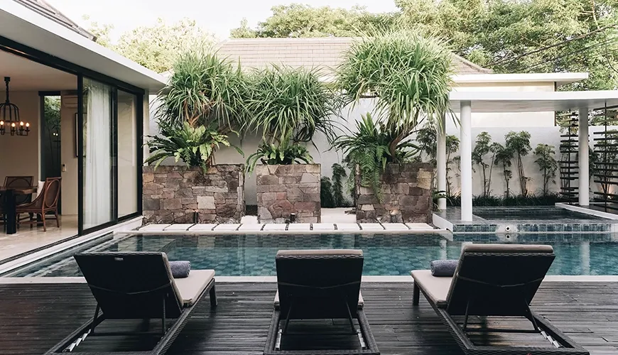 Three empty rattan lounge chairs face the refreshing swimming pool on a wooden deck surrounded by tall tropical plants and stone planters at Ambergris Villa