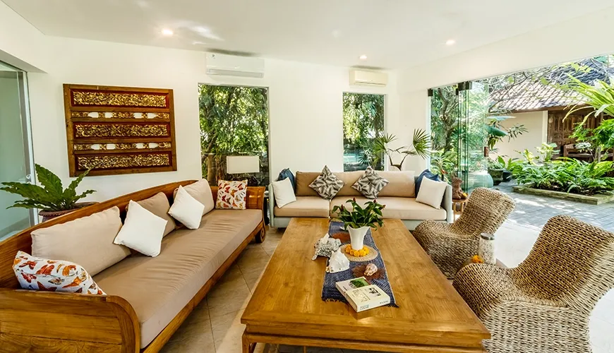 Airy villa living room featuring custom wooden sofas, woven rattan chairs, and large glass doors connecting to the garden at Asraya Villa
