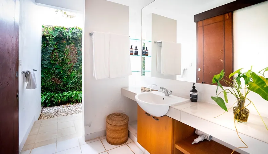 Tropical guest bathroom with a clean white vanity, large mirror, and open doorway leading to a lush outdoor garden path at Asraya Villa