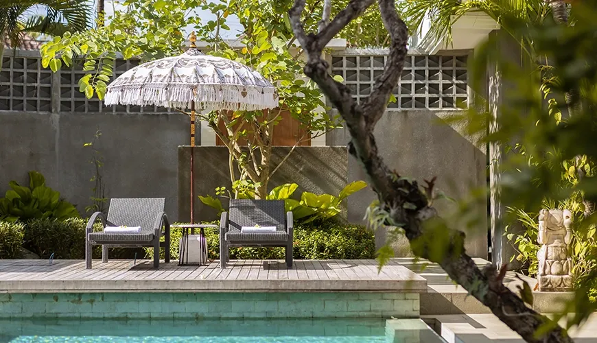 Relaxing swimming pool area with two rattan lounge chairs and a beautiful decorative parasol under lush green trees at Christie Villa