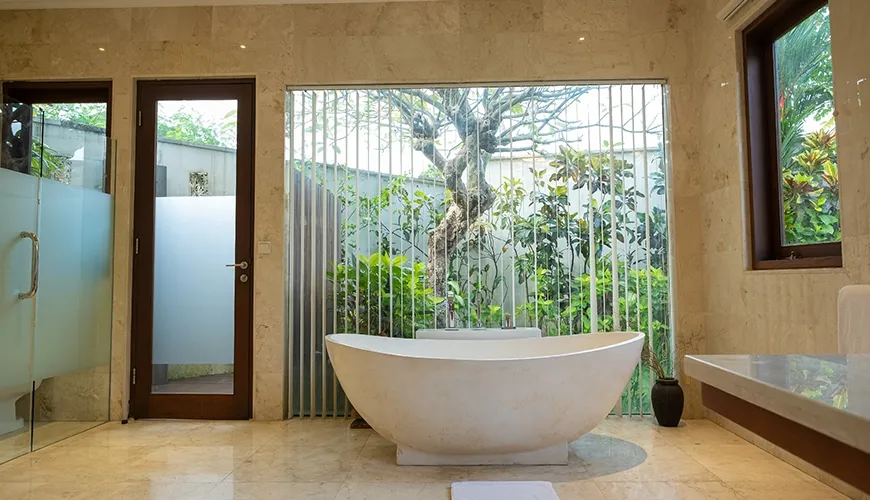 Luxurious villa bathroom featuring a beautiful freestanding bathtub, marble walls, and a large window view of a private garden at Istana Putih Villa