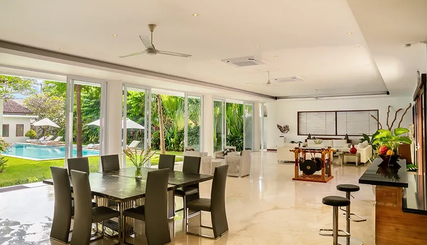 Spacious luxury villa interior with a prominent dark wood dining table, an adjacent kitchen bar, and a lounge area with pool views at Istana Putih Villa