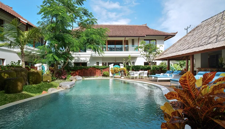 A beautiful tropical swimming pool area at a hotel with blue sun loungers and lush greenery surrounding the buildings at Kama Village 1BR