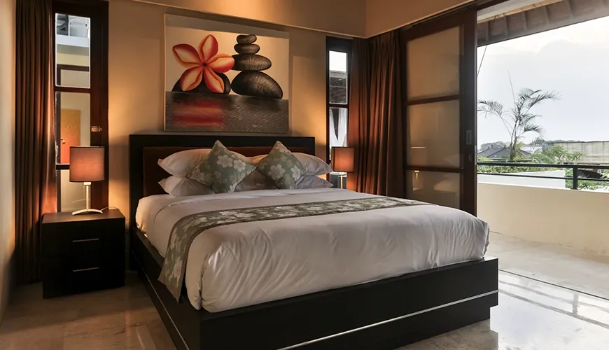 Comfortable king size bed near large sliding doors opening onto a sunny outdoor balcony with tropical foliage at Kumuda Villa