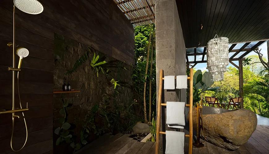 Unique tropical outdoor Maharani Bathroom featuring a rough stone bathtub, modern brass shower fixtures, and stacked towels at Le Sunset