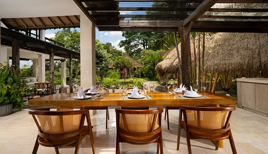 Alfresco dining area with a long wooden table and woven chairs overlooking tropical garden bungalows at Le Sunset Villa