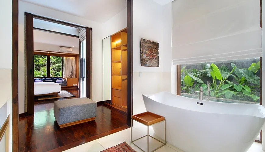 Sleek master bathroom featuring a white freestanding bathtub, dark wood floor, and large window view of tropical foliage at Leceni Villa