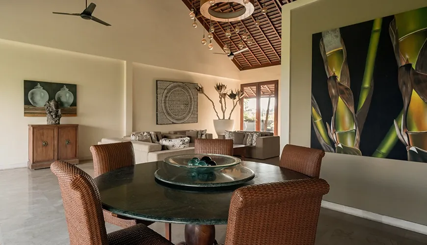 Elegant villa dining area with a beautiful marble round table, rattan chairs, contemporary artwork, and a high vaulted wooden ceiling at Omekai Cemagi