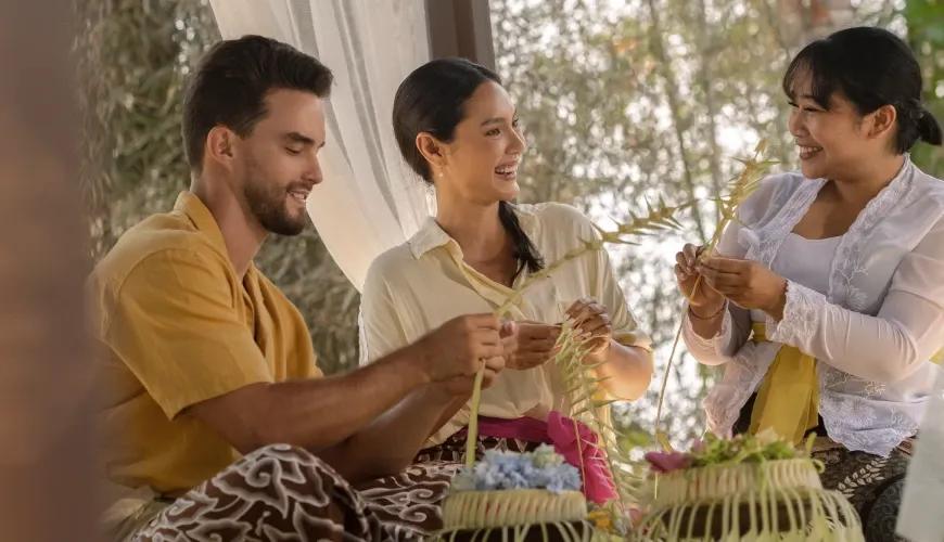 Smiling couple learning Balinese heritage and traditional crafts with villa staff in a tropical setting at Shanti Residence