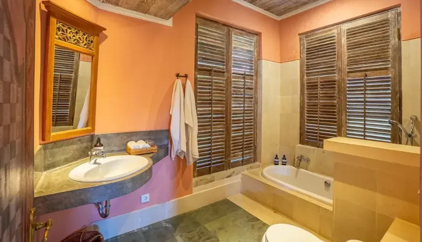 A bathroom with peach-colored walls, a large mirror, wooden shutters, and a white bathtub at Villa Angsa