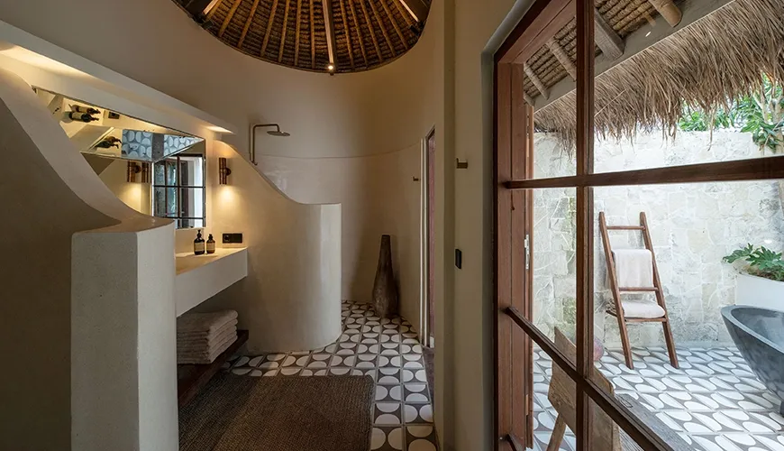 Beautiful luxury villa bathroom with a unique domed thatched roof, round mosaic tile floor, and an inviting outdoor black stone bathtub visible through the wooden doors at Villa Xanadu