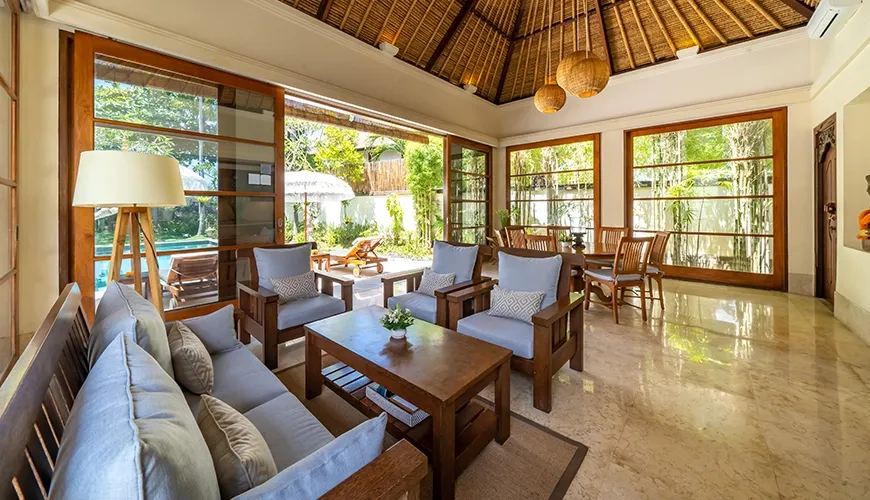 Beautiful open-plan living area of a villa with traditional wooden furniture, a thatched roof, and large windows facing the private pool at Villa Yasmine